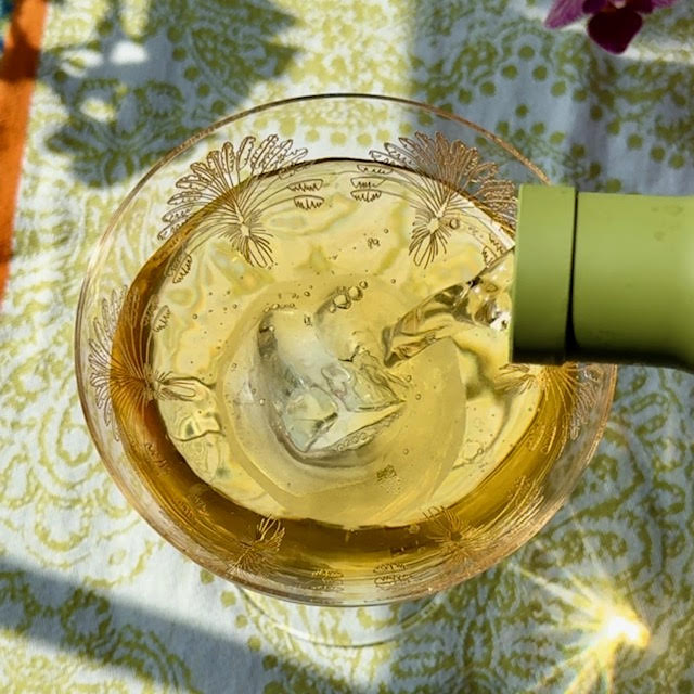 Iced green tea pouring from a green Hario Cold Brew Tea Wine Bottle into a couple glass. Sunlight shining through the glass makes a starburst pattern on the table.