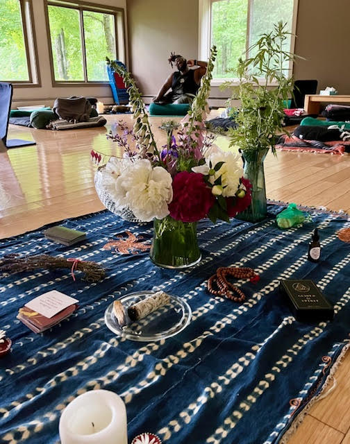 Central "altar" at a meditation training and yoga nidra retreat in 2023. Mindfulness cards, a candle, burning sage, mala beads, lavender, a bouquet of flowers, a bouquet of mugwort are in the foreground. Shawn J. Moore is in the background.