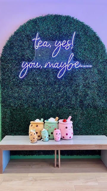 Bench with assorted bubble tea plushies in front of a neon sign that reads "tea, yes! you, maybe..." at Tea Magic in Williston Park, NY.