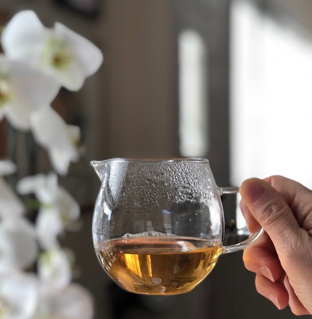 Glass fairness pitcher of amber tea with sun shining on it next to blooming white orchids.