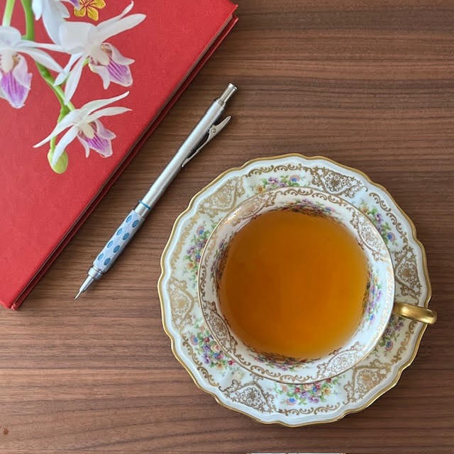 a white orchid, red journal, and mechanical pencil next to a cup of tea on a flowered saucer