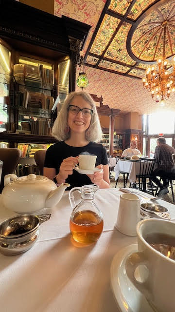 Traci AKA Tea Infusiast smiling, wearing a black shirt and glasses, while holding a steaming cup of tea at Gryphon. 