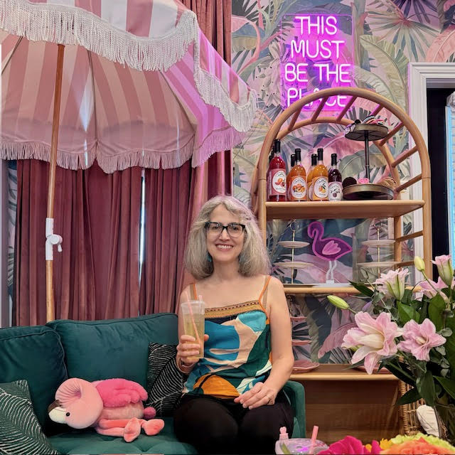 Traci AKA Tea Infusiast--a white woman with silver hair and dark green glasses smiling and holding a cup of iced tea sitting on a sofa next to a pink flamingo plushie at BesTea in Babylon, NY.