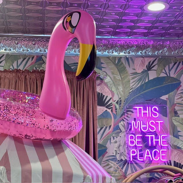 Inflatable pink flamingo pool ring on top of a pink and white umbrella next to a pink neon sign that says "THIS MUST BE THE PLACE" at BesTea in Babylon, NY.