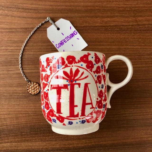 Ivory mug with the word "TEA" in red letters with a floral pattern surrounding it. A white tea-bag shaped piece of paper is attached to a tea string. The paper says "confessions."