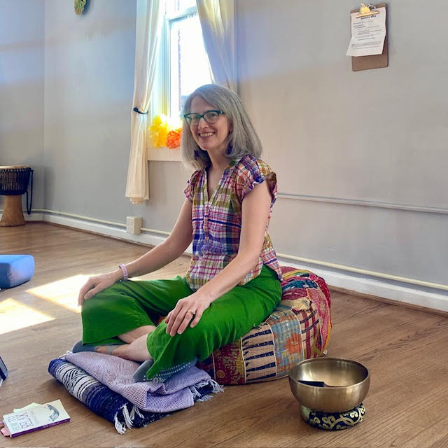 Traci Levy--a white woman with silver hair wearing a colorful shirt and green pants sitting on a yoga cushion and smiling