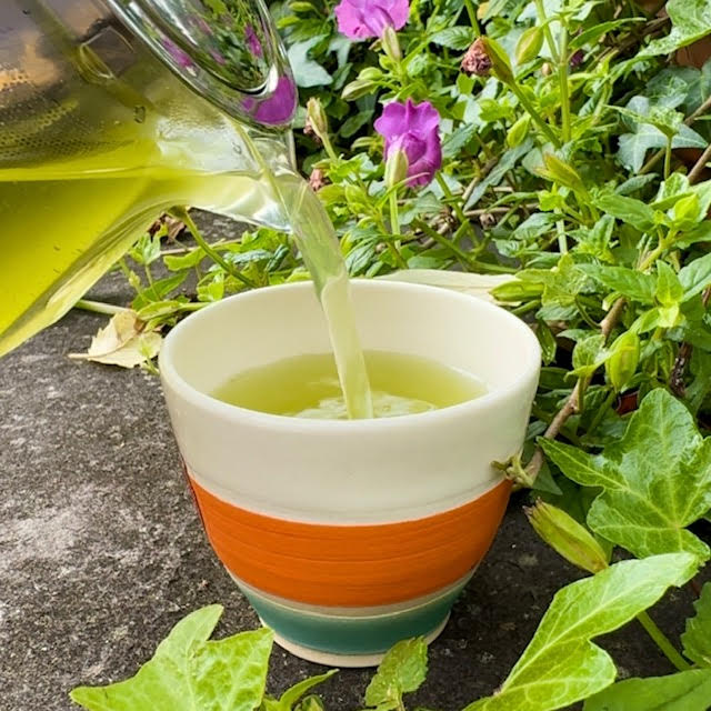 Vibrant yellow-green Yuzu Matcha Kukicha tea poured from a glass teapot into a white, orange, and blue teacup with purple flowers nearby