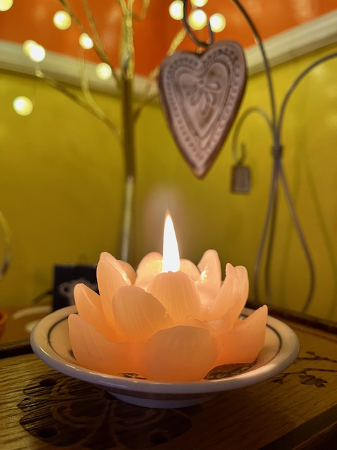 lotus shaped candle with a still flame in the foreground, tiny lights and white ceramic heart in the background