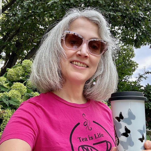 Traci Levy --AKA Tea Infusiast -- wearing sunglasses and a bright pink tea shirt that says "Tea Life" and holding a commuter mug of tea.