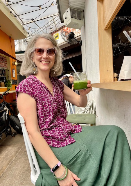 Enjoying a 2024 top tea experience: Traci Levy (AKA Tea Infusiast) holding a peach matcha shiso soda at Kettl in the Bowery. She has salt and pepper hair and is smiling while wearing sunglasses, a purple shirt, and a green skirt.