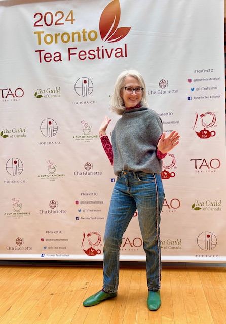 Traci Levy (AKA Tea Infusiast)--a white woman with salt and pepper hair and glasses--smiling and showing jazz hands in front of the 2024 Toronto Tea Festival sign