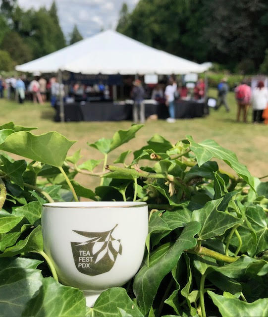 2022 TeaFest PDX white teacup with gold image and writing with an outdoor tea festival tent in the background.