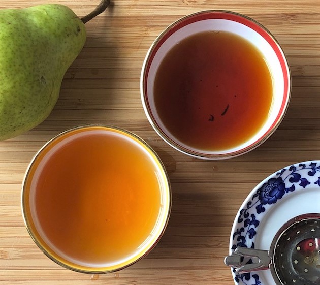 Two white teacups without handles. One has a yellow band near the top, the other a tomato orange band near the top. In the top left is a green bartlett pear. In the bottom left is a silver tea strainer on a white and blue floral saucer.