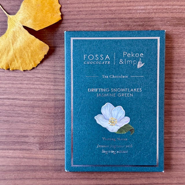 Green-tea infused chocolate bar--Drifting Snowflake Jasmine Green. The packaging is blue green, with gold writing, and a white camellia sinensis flower with gold stamens and green leaves on the front. The chocolate bar is from Fossa X Pekoe & Imp.