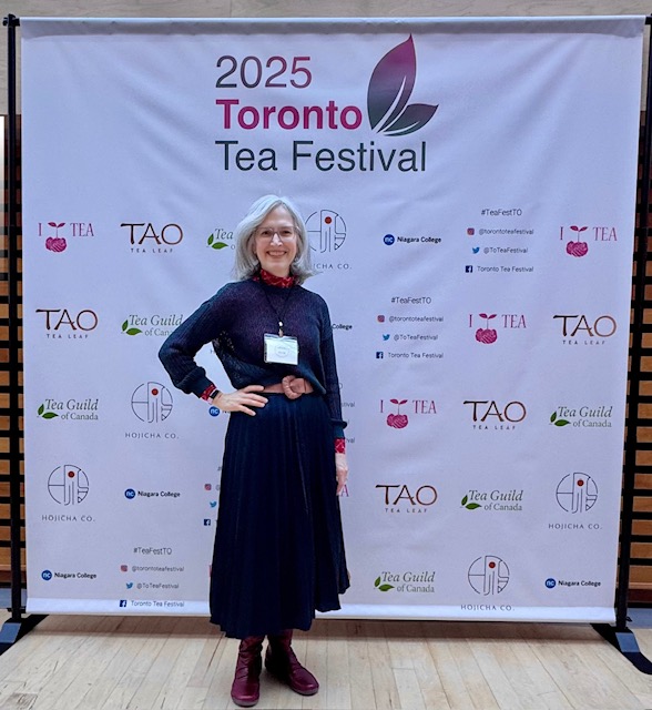 Traci Levy AKA Tea Infusiast--a white woman with should length silver hair and glasses, wearing a blue sweater and skire and red boots, standing in front of the 2025 Toronto Tea Festival sign.