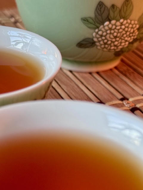 two cups of deep amber tea next to part of a green gaiwan hand painted with white hydrangeas flowers.